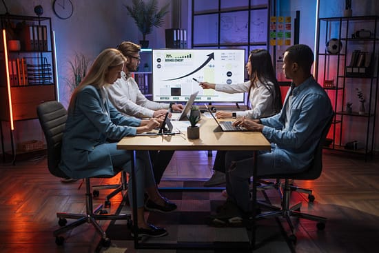 Group of diverse colleagues looking at computer monitor with worldwide business concept while sitting together at desk. Competent financiers examining graphs, charts and statistics at office.
